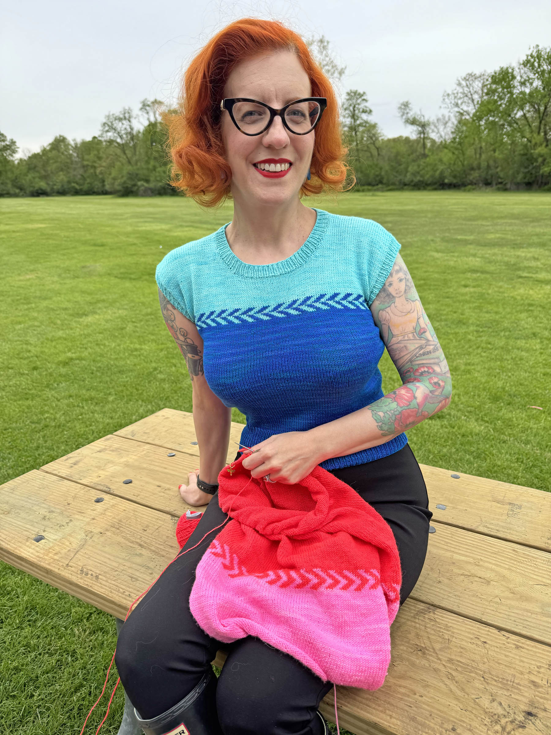 Tasha wearing an aqua and dark blue Countertone top, while knitting a pink and red version, sitting on a bench